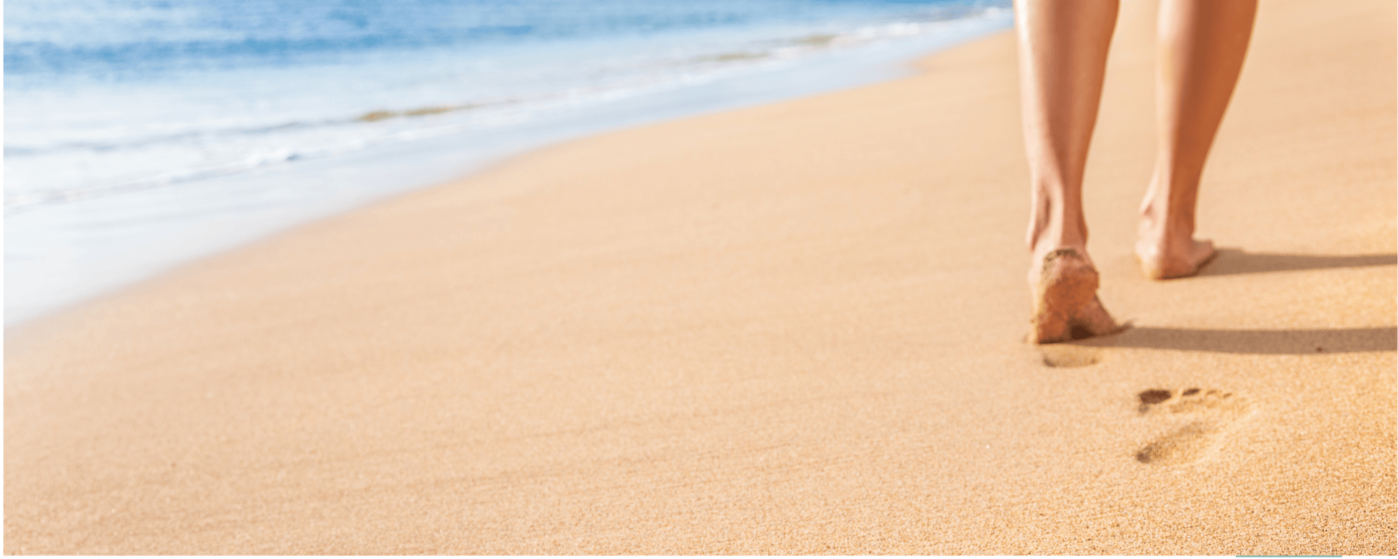 A close-up of a person's bare feet walking on a sandy beach near the ocean, with footprints in the sand.