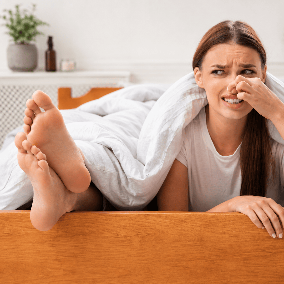 A woman lying in bed with a disgusted expression, covering her mouth with her hand, with her bare feet propped up on the wooden headboard.